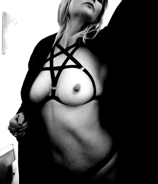 Tabitha nude with a pentagram harness highlighting breasts. Black and white. a cape is opened and thrust up to the left
